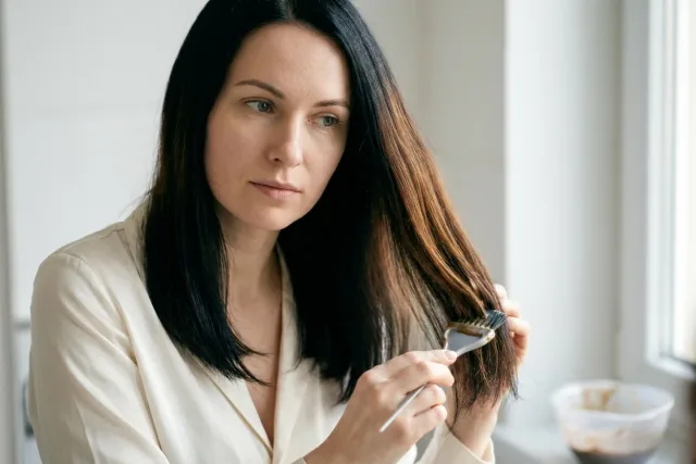 Žena si doma štětcem nanáší barvu na tmavé vlasy v koupelně.