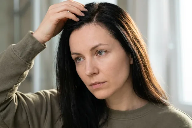 Žena v koupelně si prsty rozhrnuje černé vlasy u kořínků.