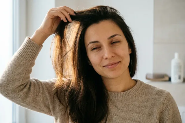Žena u okna si prsty nadzvedává dlouhé tmavě hnědé vlasy u kořínků.