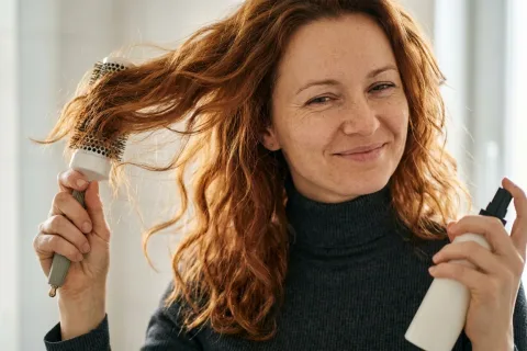 Žena fénuje vlnité vlasy a drží sprej tepelné ochrany.