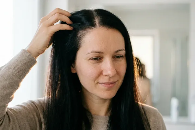 Žena u okna si prsty kontroluje pěšinku a mírně prořídnuté vlasy.